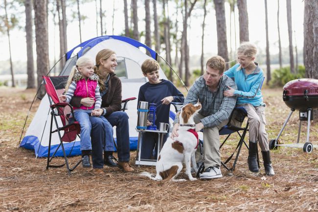 A family with three children and a dog on a camping trip in the woods, sitting by a tent, talking and smiling. The youngest child, a 5 year old girl, is sitting on mom's lap. Dad is petting the dog.